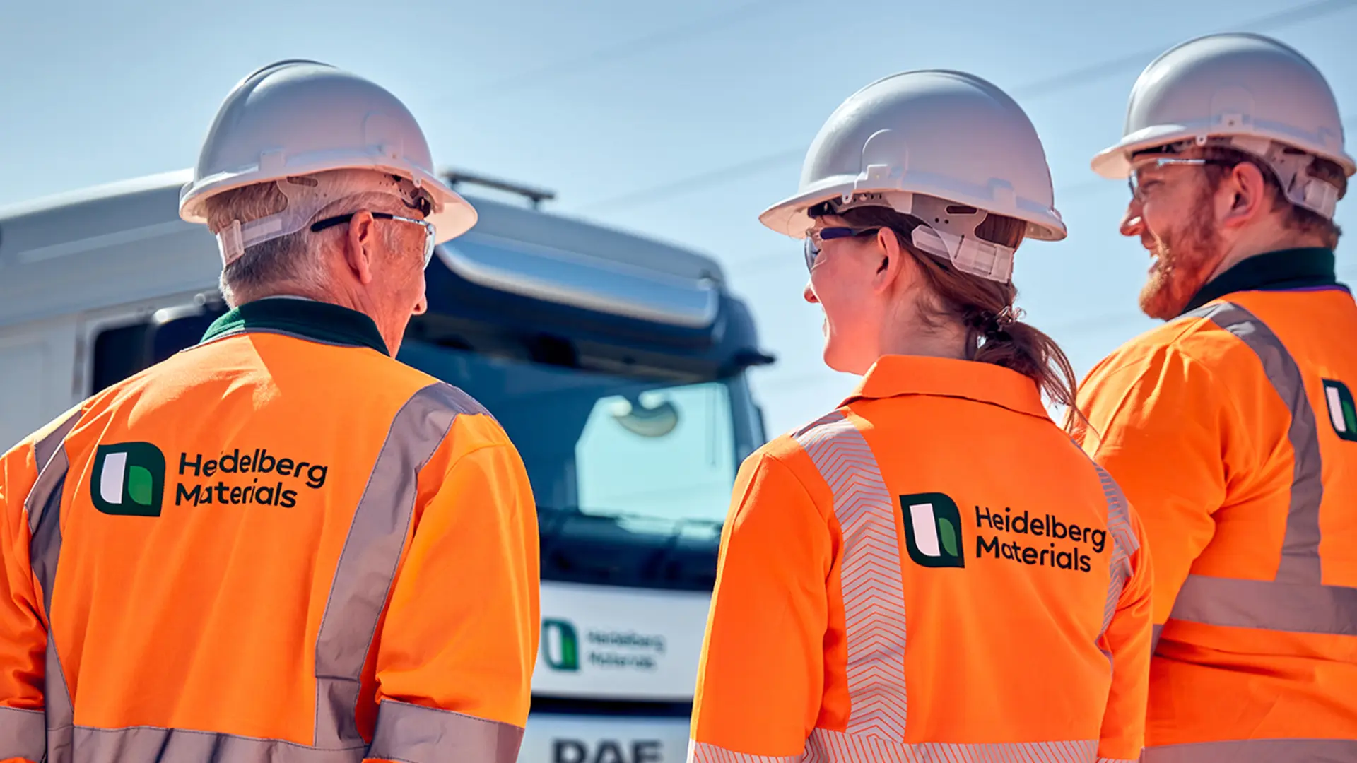 Three construction workers in high-visibility orange jackets and white helmets stand in conversation, with a logistics company logo visible on the back of their jackets. A white company truck is parked in the background under a clear blue sky.