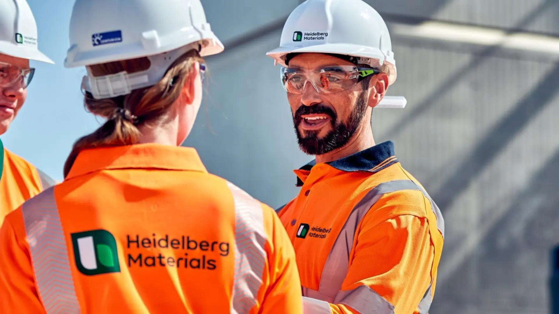 Three workers wearing orange high-visibility jackets and white safety helmets stand in conversation at a worksite, with a concrete structure and machinery in the background. The central worker's jacket displays a logo and text reading "New Zealand Hardware".