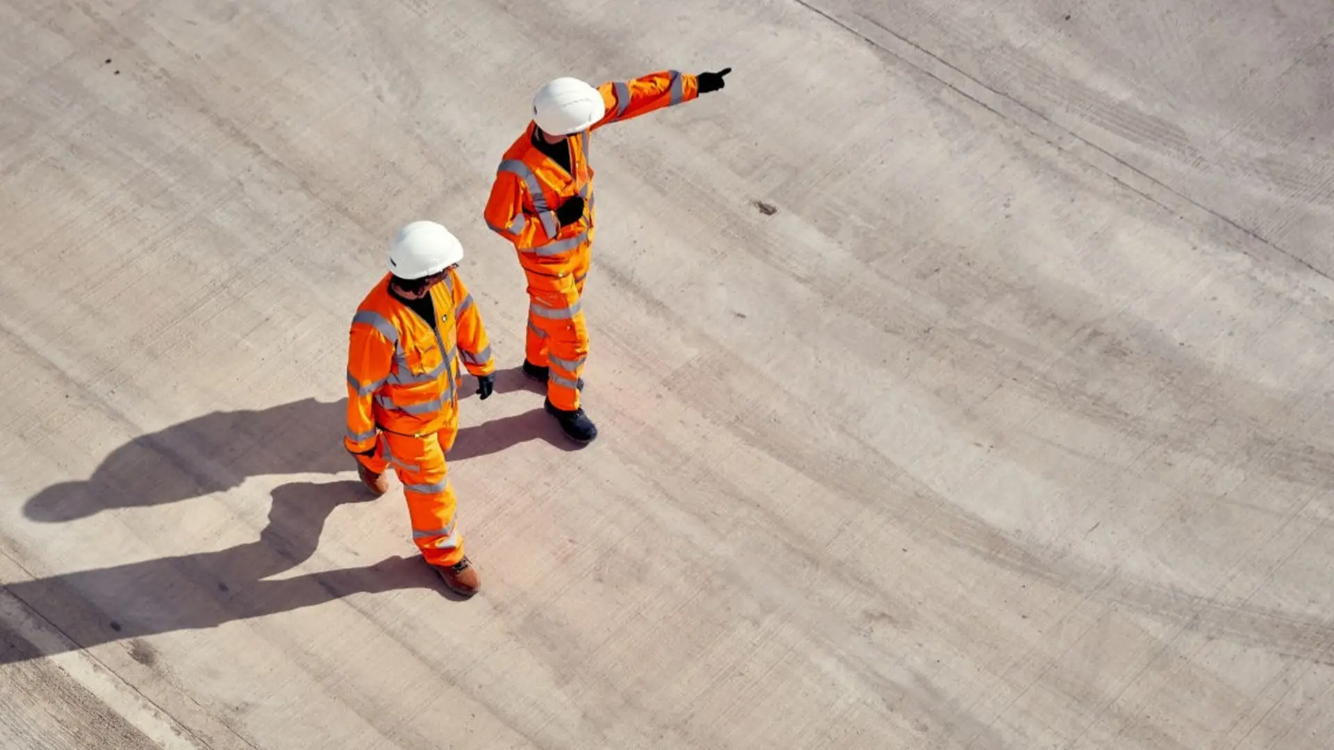 Two construction workers in orange high-visibility uniforms and white helmets stand on a large concrete surface. One worker points into the distance while the other stands beside them, facing the same direction. 