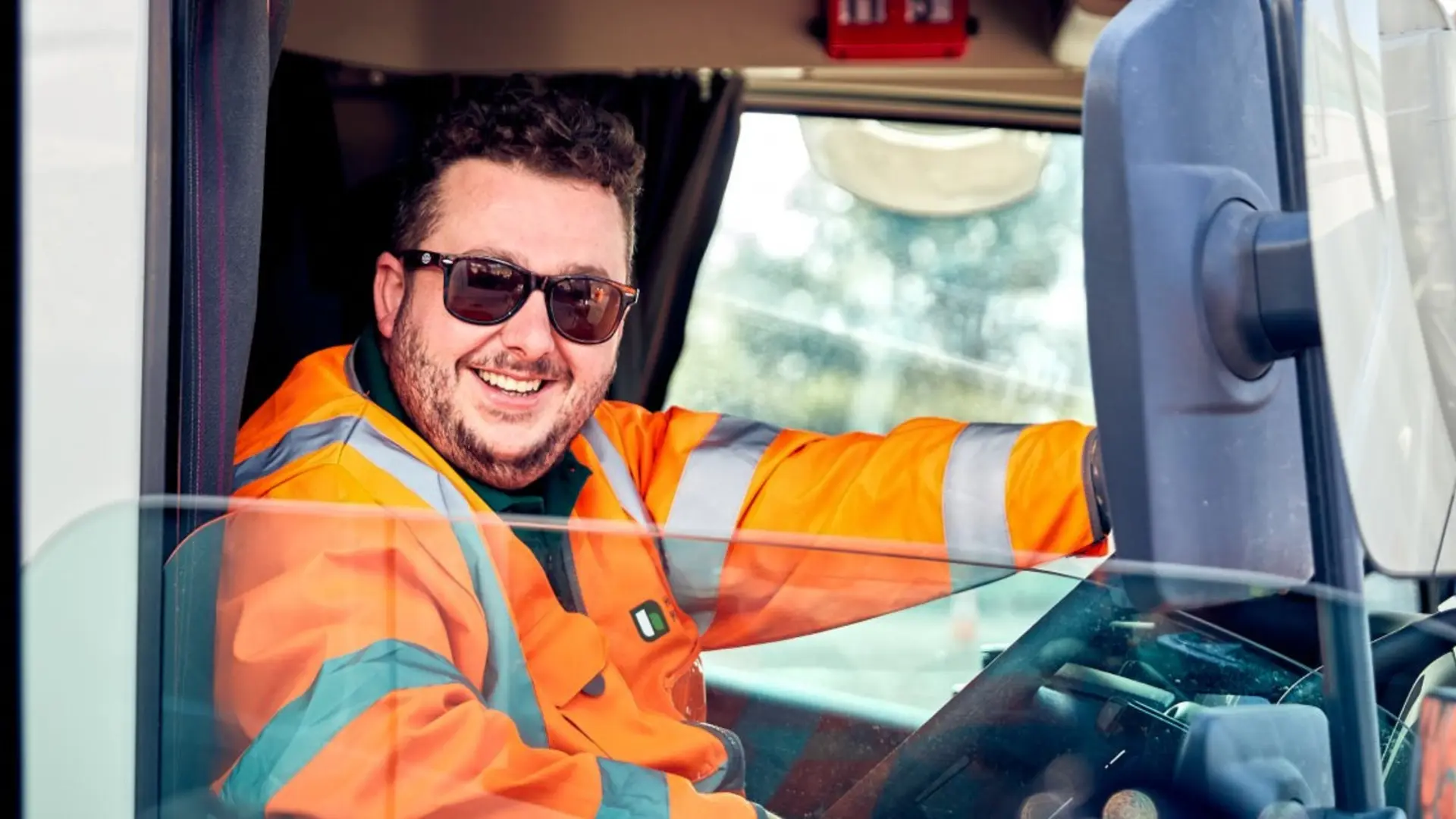 A truck driver wearing a high-visibility orange jacket sits in the driver's seat of a vehicle, holding the steering wheel with one hand. The interior of the cab and part of the side window are visible, with trees in the background outside.