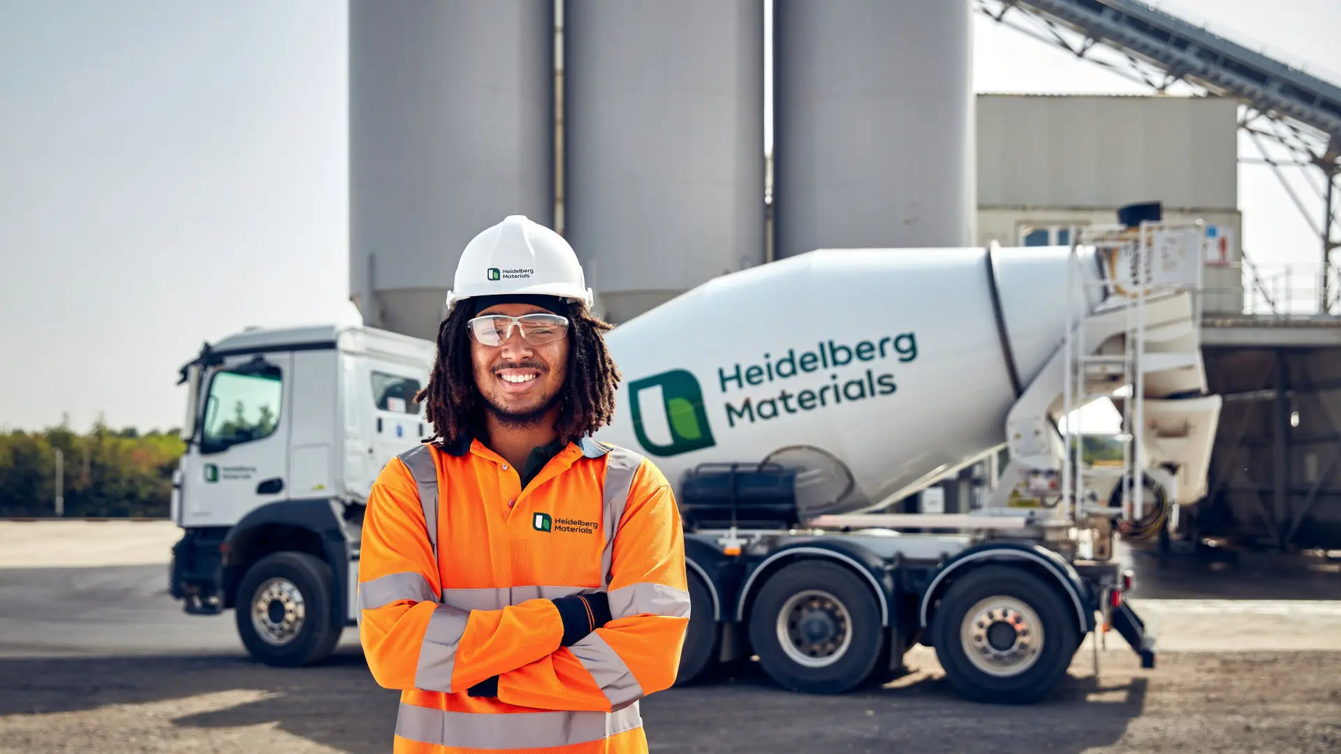 A Heidelberg Materials Concrete worker wearing a high-visibility orange jacket, safety helmet, and clear glasses stands smiling with arms crossed in front of a Heidelberg Materials branded concrete truck and large silos in an outdoor industrial setting.