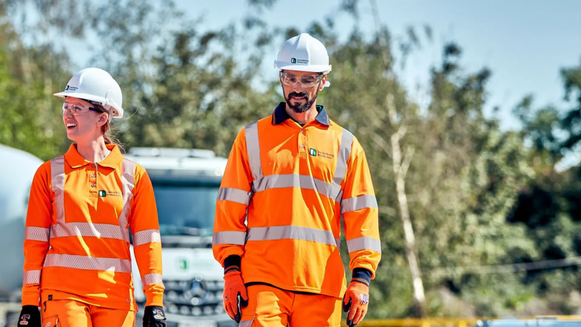 Three employees wearing orange high-visibility clothing and white hard hats walk side by side in an outdoor setting with trees in the background. A concrete mixer truck is partially visible behind them.