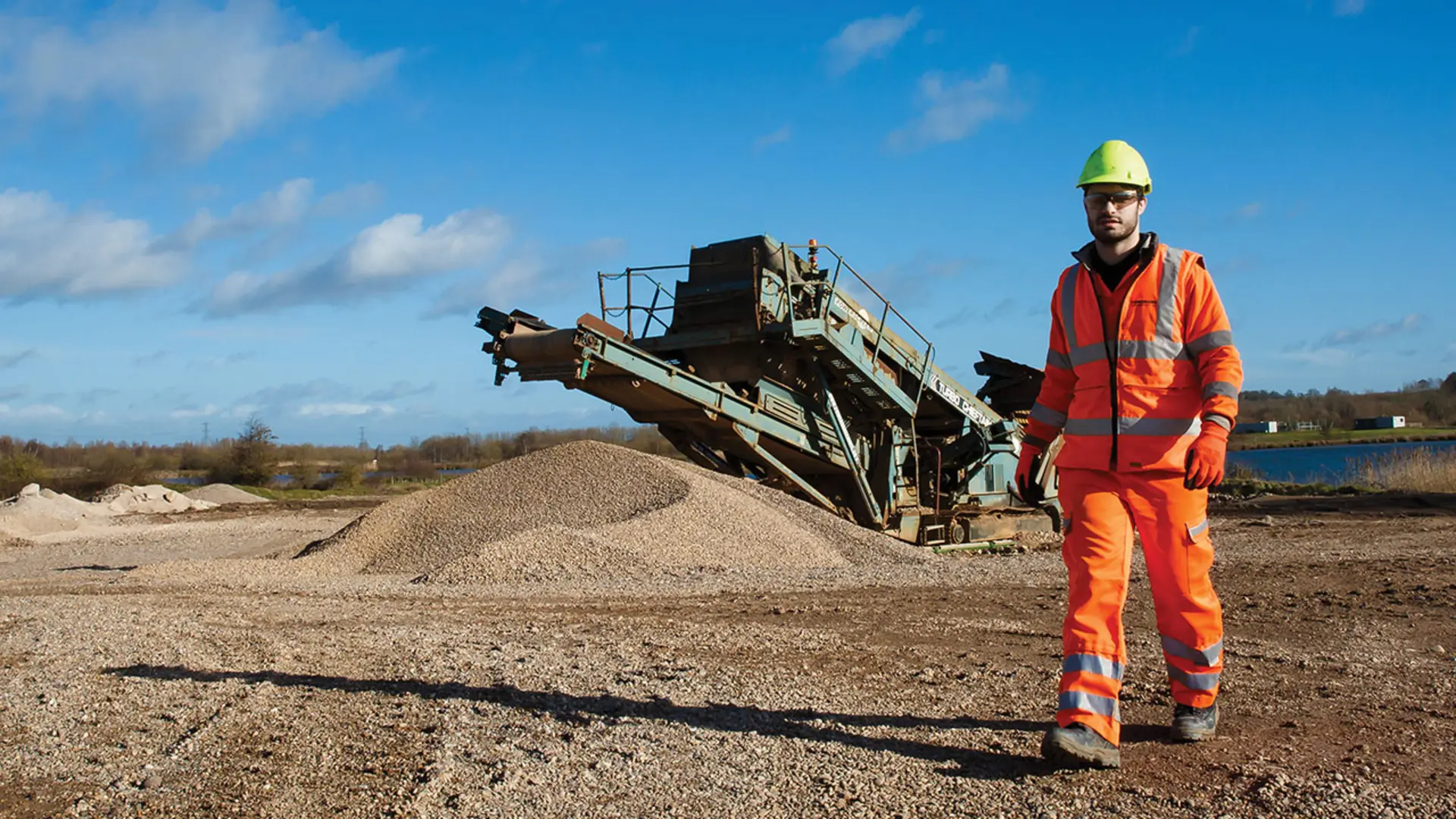 Worker in orange safety gear and helmet walking on a gravel site with a large industrial conveyor in the background, piles of gravel on the left, and a blue sky with scattered clouds above.