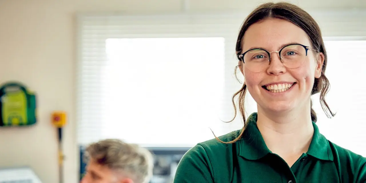 Person in a green polo shirt standing indoors with arms crossed; defibrillator mounted on the wall in the background to the left and another individual sitting at a desk partially visible in the background.