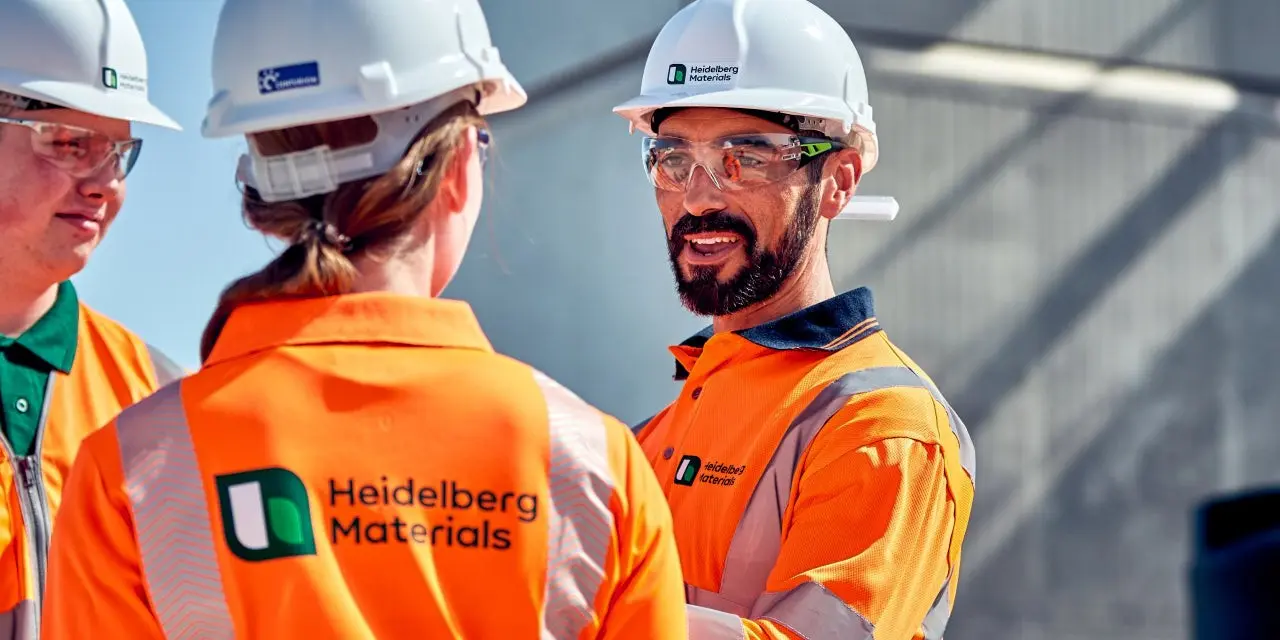 Three workers wearing orange high-visibility jackets and white safety helmets stand in conversation at a worksite, with a concrete structure and machinery in the background. The central worker's jacket displays a logo and text reading "New Zealand Hardware".