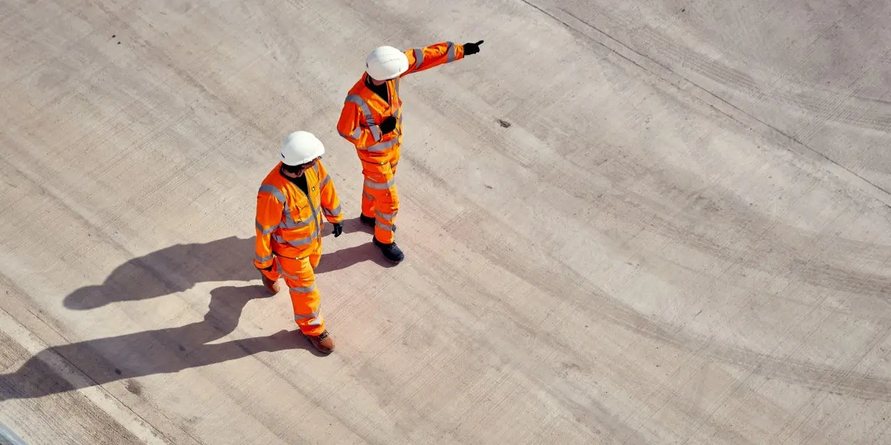 Two workers in orange safety uniforms and helmets stand on a wide concrete surface; one points into the distance while the other looks in the same direction.