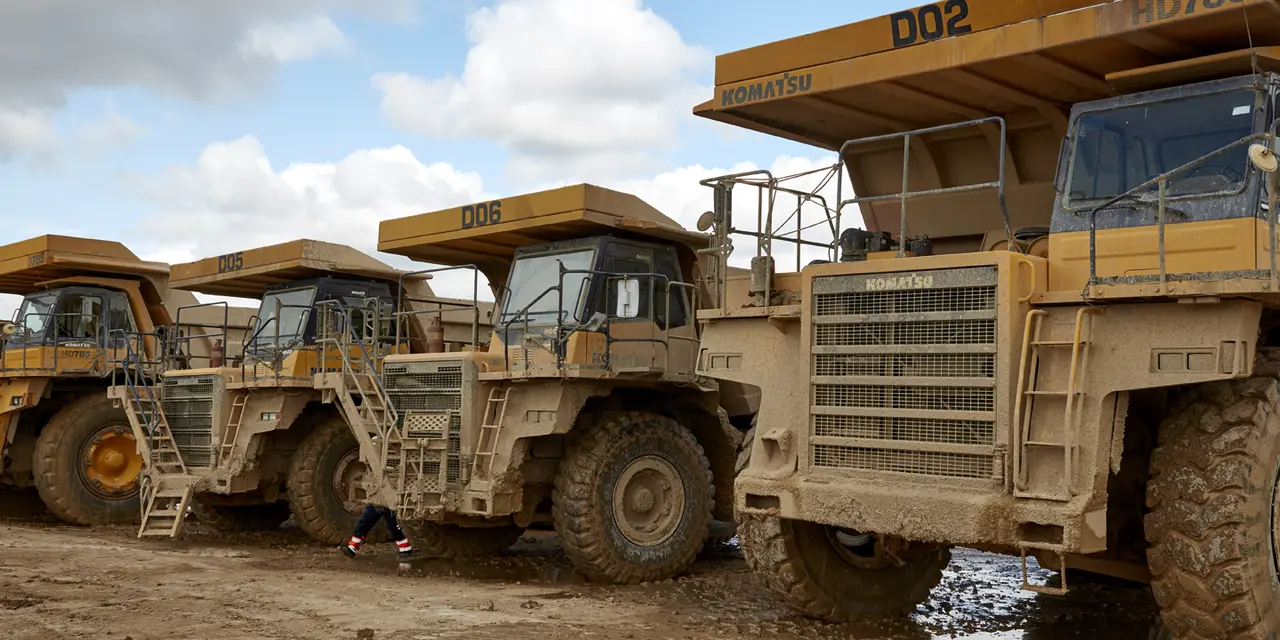 Four large yellow trucks parked in a row on a dirt lot under a cloudy sky, with visible ladders, tires, and grills, showcasing their immense size and rugged design.