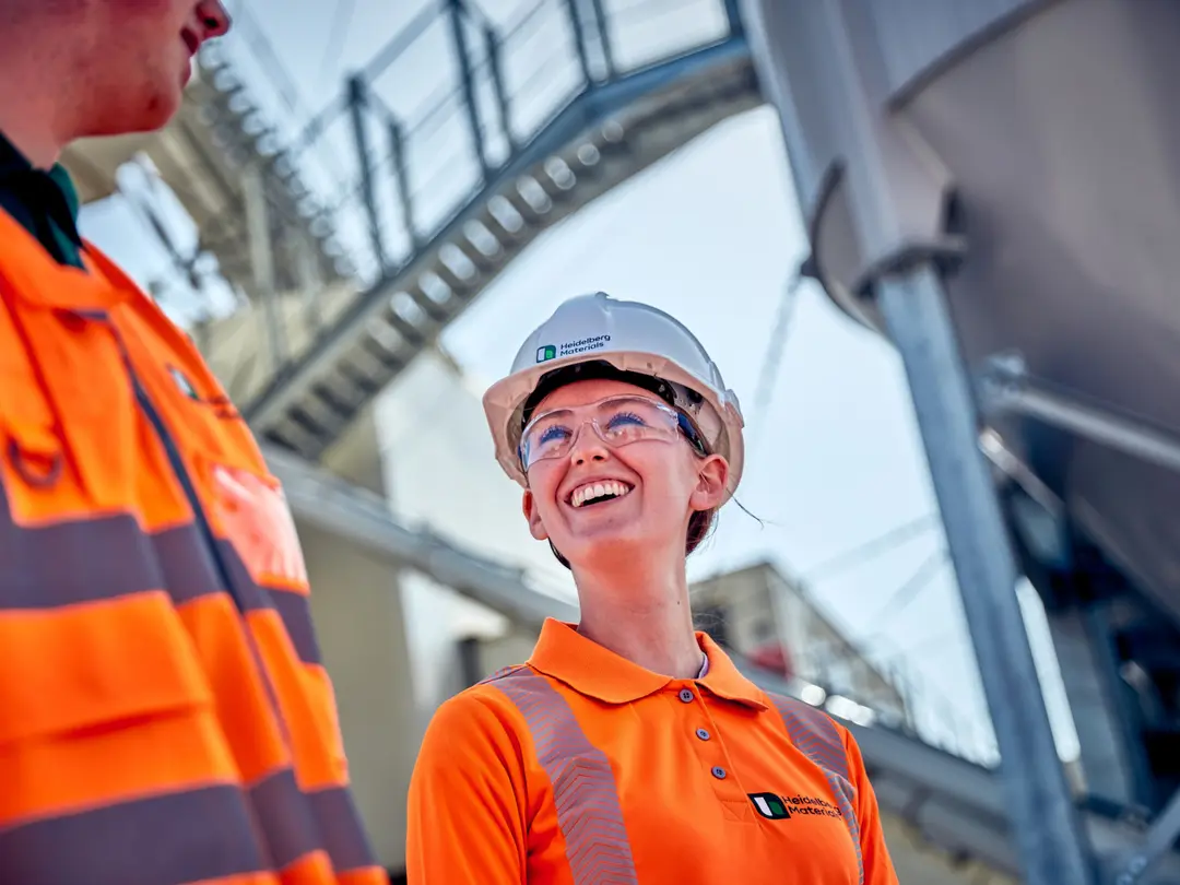 Two industrial workers wearing orange high-visibility clothing and helmets stand in an industrial site with metal structures and stairs in the background.